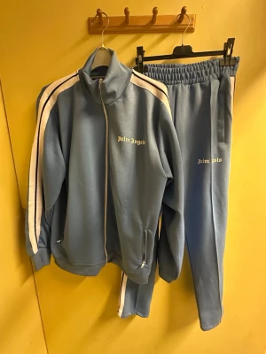 Palm Angels tracksuit.  - Säljer en blå tracksuit från Palm Angels med vit logga på benet. Byxorna har vita och svarta ränder längs sidorna, elastisk midja och raka ben. Perfekt för chill eller träning, riktigt snygga och sköna med sportig vibe.