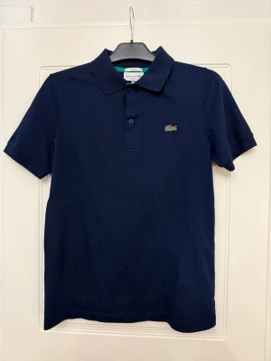 Marinblå piké från Lacoste - Snygg marinblå pikétröja från Lacoste med klassisk krage och två knappar framtill. Ikoniska krokodilloggan broderad på bröstet. Kortärmad och tillverkad i mjuk bomull som är skön mot huden. Perfekt för en clean och sportig look.