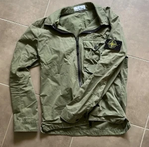Stone Island overshirt - Snygg militärgrön overshirt från Stone Island med klassisk patch på ärmen. Jackan har lång ärm, bröstficka med knapp och dragkedja framtill. Perfekt att slänga på sig för en cool streetstil. Materialet är bomull och passformen är normal. Strl: S