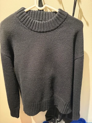 Svart stickad tröja från Monki - Mysig svart stickad tröja från Monki med grov ribbad krage, muddar vid ärmslut och nederkant. Tröjan har en enkel, klassisk design med lång ärm och relaxed fit. Perfekt för att hålla sig varm under kyliga dagar.