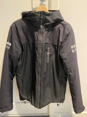 Svart Sail Racing jacka med huva - ”M Cape Insulated Jacket” från Sail Racing är en vadderad höst- och vinterjacka designad för kyliga dagar. Yttertyget består av ett tvålagers polyestermaterial som är både vind- och vattentätt samtidigt som det har bra andasförmåga. De heltejpade sömmarna gör att jackan står emot väta även under mer krävande förhållanden. Vadderingen är gjord av återvunnen polyester som ger skön värme utan att tynga ner plagget. Läs mer om Jack modellen online på SailRacing eller Stadium. Nypris 3499kr. 