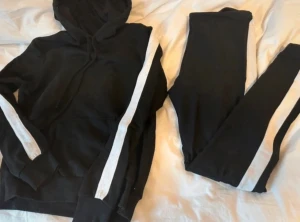 SET! Leggings med hoodie från H&M - Svart hoodie med vita ränder längs ärmarna och huva. Tröjan har snörning runt halsen och en stor magficka framtill samt med tillhörande svarta leggings med en bred vit rand längs utsidan. Sitter tight och har en sportig vibe, perfekta för en avslappnad och sportig stil