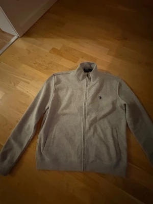 Grå zip-kofta från Polo Ralph Lauren - Snygg grå kofta från Polo Ralph Lauren med dragkedja hela vägen och klassisk ståkrage. Tröjan har lång ärm och den ikoniska Polo-loggan broderad på bröstet. Perfekt för chill dagar och enkel att matcha med jeans eller joggers.