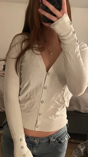 Vit kofta Zara - Supersnygg vit kofta från Zara i storlek S. Den har v-ringning, långa ärmar och är dekorerad med glittriga silverknappar både framtill och vid ärmsluten. Materialet är mjukt och tunt, perfekt för lager-på-lager. Passar dig som gillar stilrena detaljer.