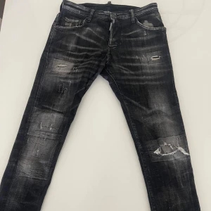 Svarta Dsquared2 jeans med slitningar - Säljer ett par svarta jeans från Dsquared2 med coola slitningar och trasiga detaljer på benen. Jeansen har en smal passform och klassisk femficksdesign. Materialet är jeans och färgen är tvättad svart med grå inslag för en snygg, använd look.