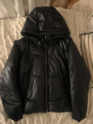 Svart pufferjacka i skinn från Zara - Säljer en svart pufferjacka från Zara i storlek S. Jackan har en snygg oversized look, huva med snörning och grov dragkedja framtill. Materialet är skinnimitation som ger en cool vibe och jackan är quiltad för extra volym. Perfekt för dig som gillar streetstyle.