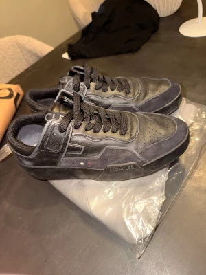 Svarta sneakers från Moncler - Snygga svarta sneakers från Moncler med rund tå och platt sula. Skorna har detaljer i skinn och mocka, samt klassisk snörning. Moncler-logga på sidan och bak på hälen. Perfekta för dig som gillar stilrena och exklusiva sneakers.