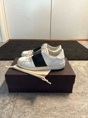 Valentino Garavani vita sneakers - Snygga vita sneakers från Valentino Garavani med bred svart rem över mitten och coola nitar på hälen. Skorna har rund tå, snörning och platt sula. Tillverkade i skinn för en lyxig känsla och stilren look. Perfekta för dig som gillar exklusiva märken och streetstyle. ÄKTA VALENTINO OPEN