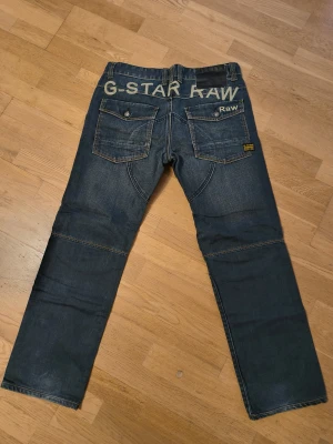 G-Star Raw R3301 jeans blå denim - Snygga blå jeans från G-Star Raw, modell R3301. De har coola detaljer med förstärkta sömmar över knäna, stora bakfickor med knapp och tydlig G-Star Raw-logga baktill. Klassisk femficksdesign och orange kontrastsömmar. Perfekt för dig som gillar streetwear.