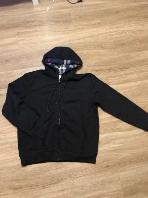 Svart hoodie med rutigt foder, stl M – Nytt skick - Snygg och stilren svart hoodie i storlek M, i helt nytt skick. Tröjan har dragkedja framtill och ett mjukt, mönstrat innerfoder som gör den både bekväm och unik i stilen. Passar perfekt till vardags, både ensam eller under en jacka.