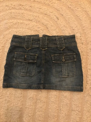 Mörkblå jeanskjol med fickor - Snygg mörkblå vintage jeanskjol med två stora fickor med knappar på baksidan och breda hällor i midjan. Kjolen har klassisk denimlook och stängs med dragkedja och knapp framtill. 