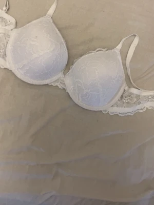 Vit spets-bh 75B med blommönster - Snygg vit bh i storlek 75B med blommig spets och tunna axelband. Materialet är 90% polyamid och 10% elastan, vilket gör den mjuk och följsam. Kuporna har ett diskret blommönster och spetsdetaljer längs kanterna för en extra feminin look.
