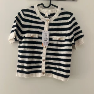 Randig kofta med kort ärm från Gina Tricot - Snygg kofta från Gina Tricot med svart och vit randigt mönster, korta ärmar och guldfärgade knappar framtill. Två dekorativa fickor med knappdetaljer på bröstet. Mjuk stickad kvalitet och ribbade kanter för extra stil.