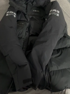 Svart dunjacka Sail Racing - Svart dunjacka från Sail Racing med huva och feta loggor på ärmarna och bröstet. Jackan har lång ärm, tvåvägsdragkedja och är rejält puffad för max värme. Materialet är syntet och dun, perfekt när det är kallt ute. Snygg och stilren design med mörk ton.