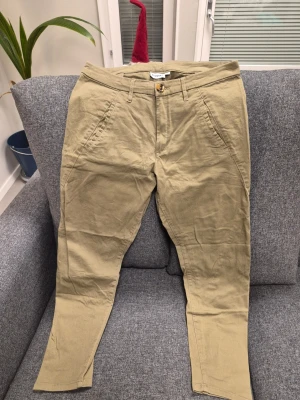 Olivgrön chinos från Kappahl, storlek 44 - Snygga beige chinos från Kappahl i storlek 44. Byxorna har normal passform, raka ben och klassiska fickor fram och bak. Tillverkade i mjuk bomull med bälteshällor och en cool knappdetalj i midjan. Perfekta för en chill och stilren look.