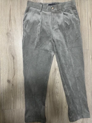 Grå manchesterbyxor från Pinque - Snygga grå manchesterbyxor från Pinque med klassisk rak passform och diskreta fickor både fram och bak. Byxorna har bälteshällor och stängs med knapp och dragkedja. Perfekta för dig som gillar stilrena och mjuka material.