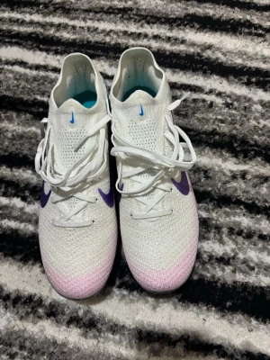 Nike Mercurial fotbollsskor vit/lila - Nike Mercurial fotbollsskor i vit med lila och blå detaljer. Skorna har snörning, rund tå och en platt sula med blå och lila dobbar. Ovandelen är stickad och har en gradient från vitt till ljusrosa framtill. Snyggt Nike-märke i lila på sidan och blå insida.