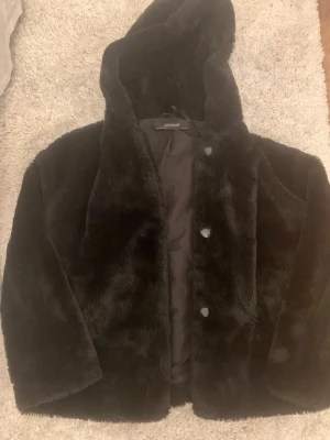 Svart pälsjacka med huva från Zara - Mjuk och fluffig svart pälsjacka från Zara Basic med huva och knappar framtill. Jackan har en loose passform och är perfekt för kalla dagar. Materialet är syntetpäls som känns riktigt gosigt och ger en cool look. Snygg att styla med jeans eller sneakers.