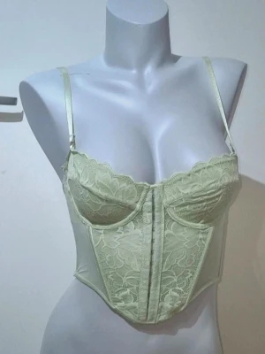 Ljusgrön spetskorsett med justerbara band - Säljer en ljusgrön korsett med tunna, justerbara axelband och vacker spets framtill. Toppen har hakknäppning bak och detaljerade sömmar som ger en snygg siluett. Perfekt för dig som vill ha en feminin och trendig look. Storlek 70C