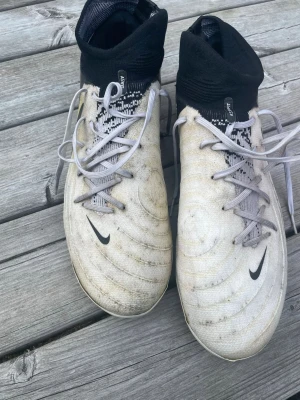 Nike fotbollsskor Flyknit strl 46 - Säljer ett par grymma Nike fotbollsskor med Flyknit-teknologi i storlek 46. Skorna är vita med svarta och guldiga detaljer, har snörning och en strumpliknande svart krage för extra stöd. Snyggt mönster på ovandelen och platt sula med dobbar för bra grepp på planen.