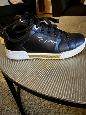Svarta sneakers från Tommy Hilfiger - Snygga svarta sneakers från Tommy Hilfiger med gulddetaljer och vit sula. Skorna har snörning och är tillverkade i skinn med märkets logga i guld på sidan. Perfekta för dig som gillar stilrena och trendiga skor med exklusiv känsla.