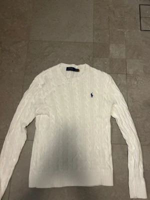 Vit kabelstickad tröja Polo Ralph Lauren - Snygg vit kabelstickad tröja från Polo Ralph Lauren med klassisk blå logga broderad på bröstet. Rund hals, ribbade muddar och långärmad. Perfekt för dig som gillar stilrena och tidlösa plagg med premiumkänsla. Priset är inte hugget i sten och kan diskuteras vid en snabb affär!! Köpt ny från care of Carl för 1599kr knappt använd!!