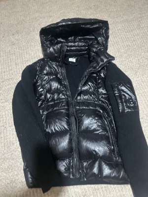 Svart pufferjacka från C.P. Company - Svart pufferjacka från C.P. Company med glansig polyester på framsidan och huvan, samt stickade ärmar och rygg. Jackan har dragkedja framtill och en cool detalj med logotyp på ärmen. Perfekt för dig som vill ha en unik och snygg jacka.