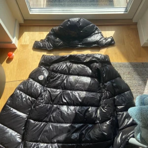 Svart dunväst från Moncler med huva - Svart dunväst från Moncler med glansig finish, huva och dragkedja framtill. Västen har två sidofickor med dragkedja, Moncler-logga på bröstet och coolt serietryck på insidan. Perfekt för lager-på-lager och streetstyle. Fylld med vitt gåsdun för extra värme.