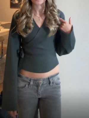 Mörkgrön omlott kofta med knyt - Säljer en mörkgrön omlott kofta med knyt i sidan och breda, långa ärmar. Tröjan är croppad och har ribbad nederkant som ger en snygg passform. Perfekt för dig som gillar trendiga och bekväma plagg.