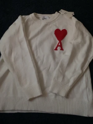 Vit stickad tröja från Ami - Vit stickad tröja från Ami med rund hals och ribbade muddar. På bröstet finns ett rött hjärta och bokstaven A som broderad detalj. Tröjan har lång ärm och en clean, klassisk look som är lätt att matcha med jeans eller chinos.
