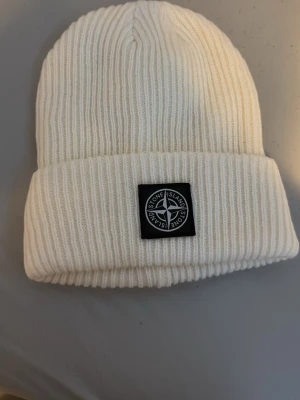 Vit ribbstickad mössa från Stone Island - Säljer en vit ribbstickad mössa från Stone Island med klassisk logopatch framtill. Mössan har uppvikt kant och är tillverkad i mjukt stickat material som håller dig varm och snygg under kyliga dagar.