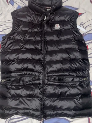 Svart dunväst från Moncler - Snygg svart dunväst från Moncler med hög krage och två fickor med dragkedja framtill. Västen har quiltad design och klassisk Moncler-logga på bröstet. Perfekt för lager-på-lager och håller dig varm under kyliga dagar.