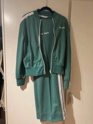 Palm Angels gröna mjukisbyxor - Snygga gröna mjukisbyxor från Palm Angels med vita och svarta ränder längs sidorna. Byxorna har en loose passform och är tillverkade i polyester. Loggan är tryckt i vitt framtill. Perfekt för chill eller streetwear.