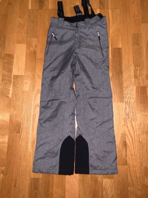 Grå skidbyxor från Five Seasons - Säljer ett par grå skidbyxor från Five Seasons med svarta förstärkningar vid bensluten och praktiska hängslen. Byxorna har dragkedjor på fickorna och är gjorda i slitstarkt syntetmaterial, perfekta för kalla dagar utomhus och aktiviteter som skid samt snowboardåkning 