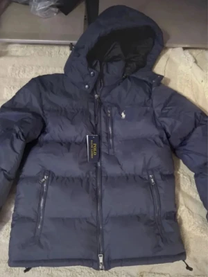Mörkblå dunjacka från Polo Ralph Lauren - Säljer en mörkblå dunjacka från Polo Ralph Lauren med huva och två sidofickor med dragkedja. Jackan har ett klassiskt puffer-utseende och Polo-loggan på bröstet. Perfekt för kalla dagar och har en snygg, sportig vibe.