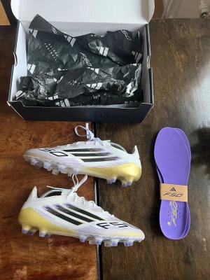 Adidas F50 fotbollsskor vit/guld - NYSKICK - Säljer ett par Adidas F50 fotbollsskor i vitt med svarta och lila detaljer samt guld på hälen. Skorna har snörning, platt sula med dobbar och kommer med lila innersulor. Perfekt för dig som vill ha grymma skor på planen. Nyskick med kvitto!