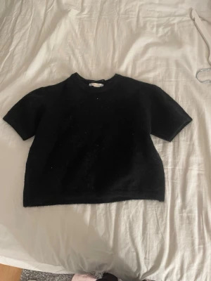 Svart stickad kortärmad topp - En svart stickad topp med korta ärmar och rund hals. Modellen är lite croppad och har en enkel, stilren design utan synliga detaljer eller mönster. Perfekt att matcha med jeans eller kjol för en chill look.
