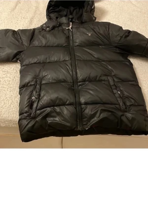 Svart pufferjacka från Gant - Säljer en svart pufferjacka från Gant med huva och dragkedja framtill. Jackan har två fickor med knappstängning och en insida i rött och grått tyg. Storlek 170-180.