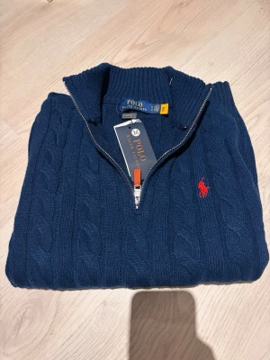 stickad tröja 1/4 zip Polo Ralph Lauren - Säljer en marinblå stickad tröja från Polo Ralph Lauren med klassiskt kabelmönster och röd broderad logga på bröstet. Tröjan har lång ärm, ribbad krage och half zip-dragkedja framtill. Perfekt för dig som gillar stilren och tidlös design. Är i M men passar bara S. Dsvdv. Skriv t mig för frågor osv. 