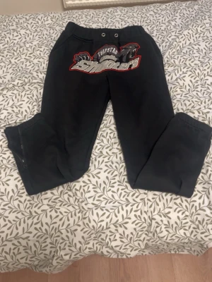 Trapstar shooters mjukisbyxor svart S - Svarta mjukisbyxor från Trapstar med stort broderat shooters-motiv i grått, vitt och rött framtill. Byxorna har snörning i midjan, dragkedjor vid bensluten och är gjorda i mjukt bomullsmaterial. Perfekt för chill och streetwear.