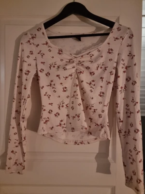 Blommig långärmad topp med snörning - Säljer en ljus, ribbad långärmad topp med små röda blommor och v-ringad hals. Toppen har en snygg snörningsdetalj framtill och spetskant vid halsen. Materialet känns mjukt och stretchigt, perfekt för vår och sommar. Passformen är normal och toppen är croppad.