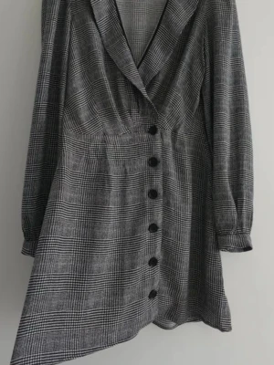 Rutig kortklänning från Reformation - Snygg kortklänning från Reformation i svart och grått rutigt mönster. Klänningen har omlott-design med djup v-ringning, långa ärmar och knappar framtill. Den har även ett knytband i midjan för extra passform. Perfekt för dig som gillar trendiga och klassiska plagg.