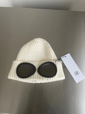 Vit C.P. Company Mössa - Vit ribbstickad mössa från C.P. Company med ikoniska svarta glasögonlinser framtill. Mössan är tillverkad i mjukt material och har en uppvikt kant för extra stilpoäng. Perfekt accessoar för att sticka ut i mängden.