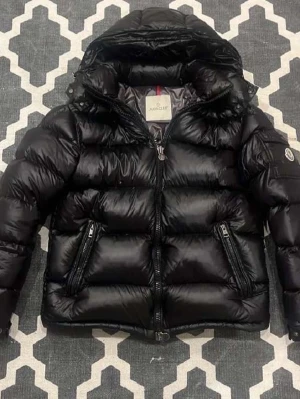Svart pufferjacka från Moncler - Säljer en svart pufferjacka från Moncler med glansig finish och huva. Jackan har två snedställda fickor med dragkedja, Moncler-logga på ärmen och dubbla dragkedjor framtill. Perfekt för dig som vill ha en snygg och varm jacka med streetkänsla.