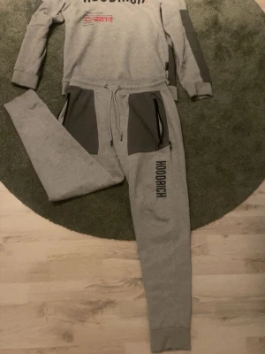 Grå mjukisbyxor från Hoodrich - Snygga grå mjukisbyxor från Hoodrich med mörkgrå paneler vid fickorna och svart logotyptryck längs benet. Byxorna har snörning i midjan och dragkedjor på fickorna för extra funktion. Perfekta för chill eller streetwear-stil.