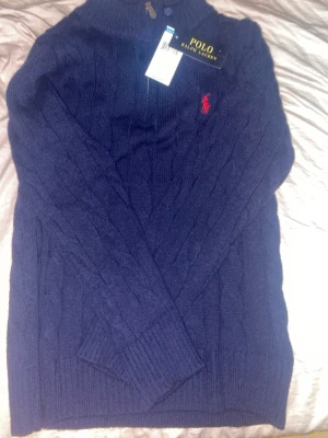 Mörkblå stickad tröja Polo Ralph Lauren - Säljer en mörkblå stickad tröja från Polo Ralph Lauren i storlek S. Tröjan har kabelstickat mönster, lång ärm och en liten röd logga broderad på bröstet. Ribbad vid hals, ärmslut och nederkant. Perfekt för kyliga dagar.