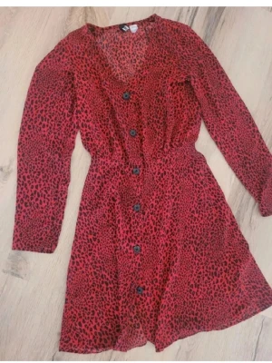 Röd leopardmönstrad klänning från h&m - Snygg röd kortklänning från h&m divided med svart leopardmönster, v-ringad hals och knappar framtill. Klänningen har långärmade ärmar och markerad midja för en smickrande siluett. Perfekt för dig som gillar att sticka ut med djurmönster. Stl 34.