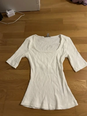 Vit ribbad topp från H&M - Säljer en vit ribbad topp från H&M i storlek XS. Toppen har en fyrkantig urringning och trekvartsärm, vilket ger en snygg och modern look. Materialet är mjukt och stretchigt, perfekt för att matcha med jeans eller kjol.