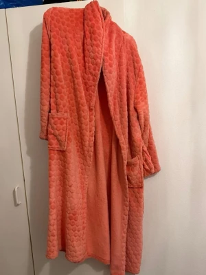 Korallfärgad morgonrock  - Säljer en mysig korallfärgad morgonrock med prickigt mönster. Västen har två fickor framtill och en bred krage som ger extra komfort. Perfekt för att hålla sig varm och chill hemma eller när du vill ha något mjukt på dig.