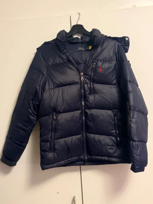 Mörkblå pufferjacka från Polo Ralph Lauren - Snygg mörkblå pufferjacka från Polo Ralph Lauren med huva och dragkedja framtill. Jackan har två sidofickor med dragkedja och ett rött Polo-logo broderat på bröstet. Perfekt för kalla dagar och har en schysst, quiltad look.